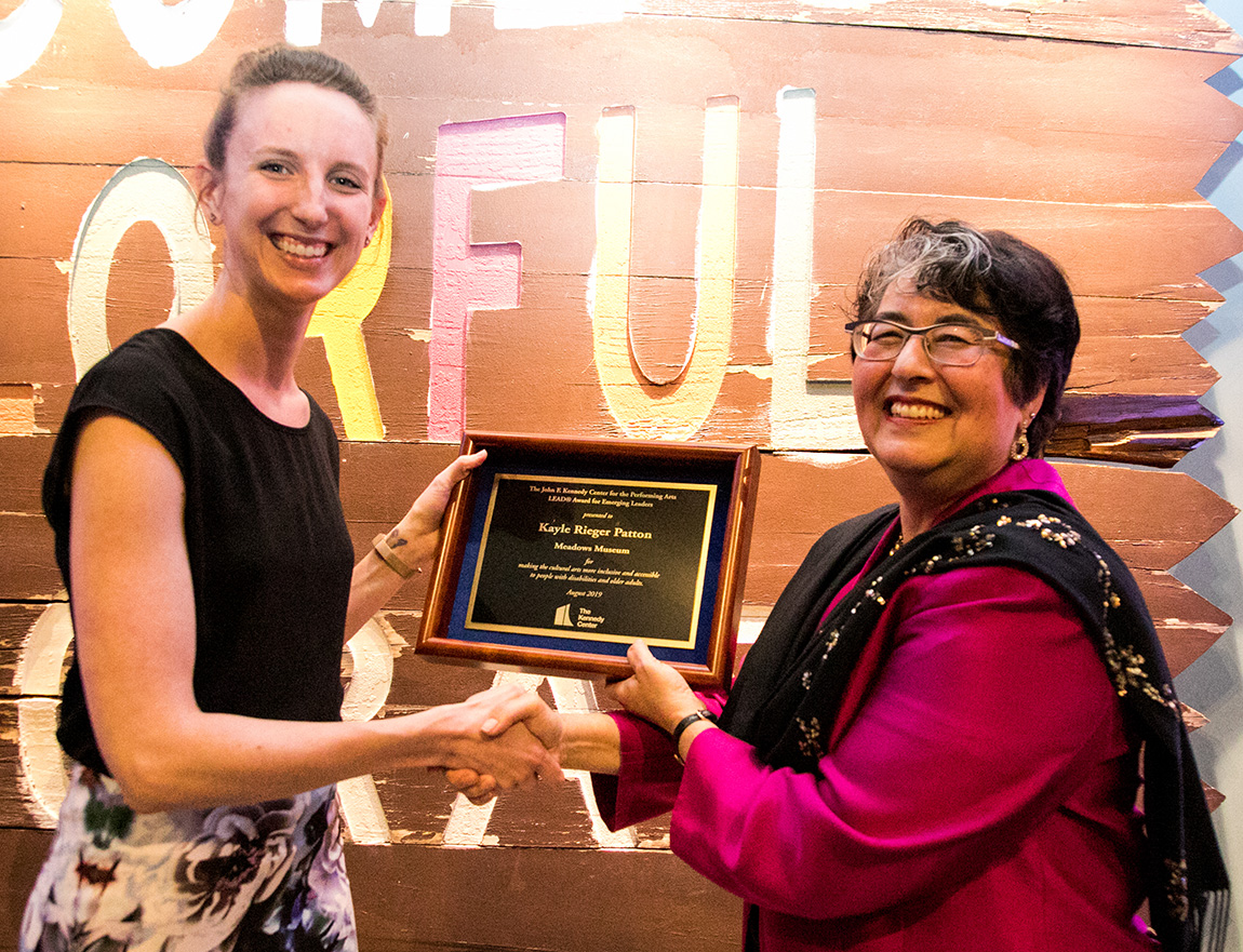 A tall woman shaking hands with a shorter woman while being presented with a framed award