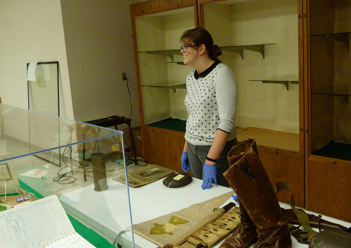 A woman changing out a museum exhibit, with empty glass shelves behind her and artifacts sitting on a table in front of her