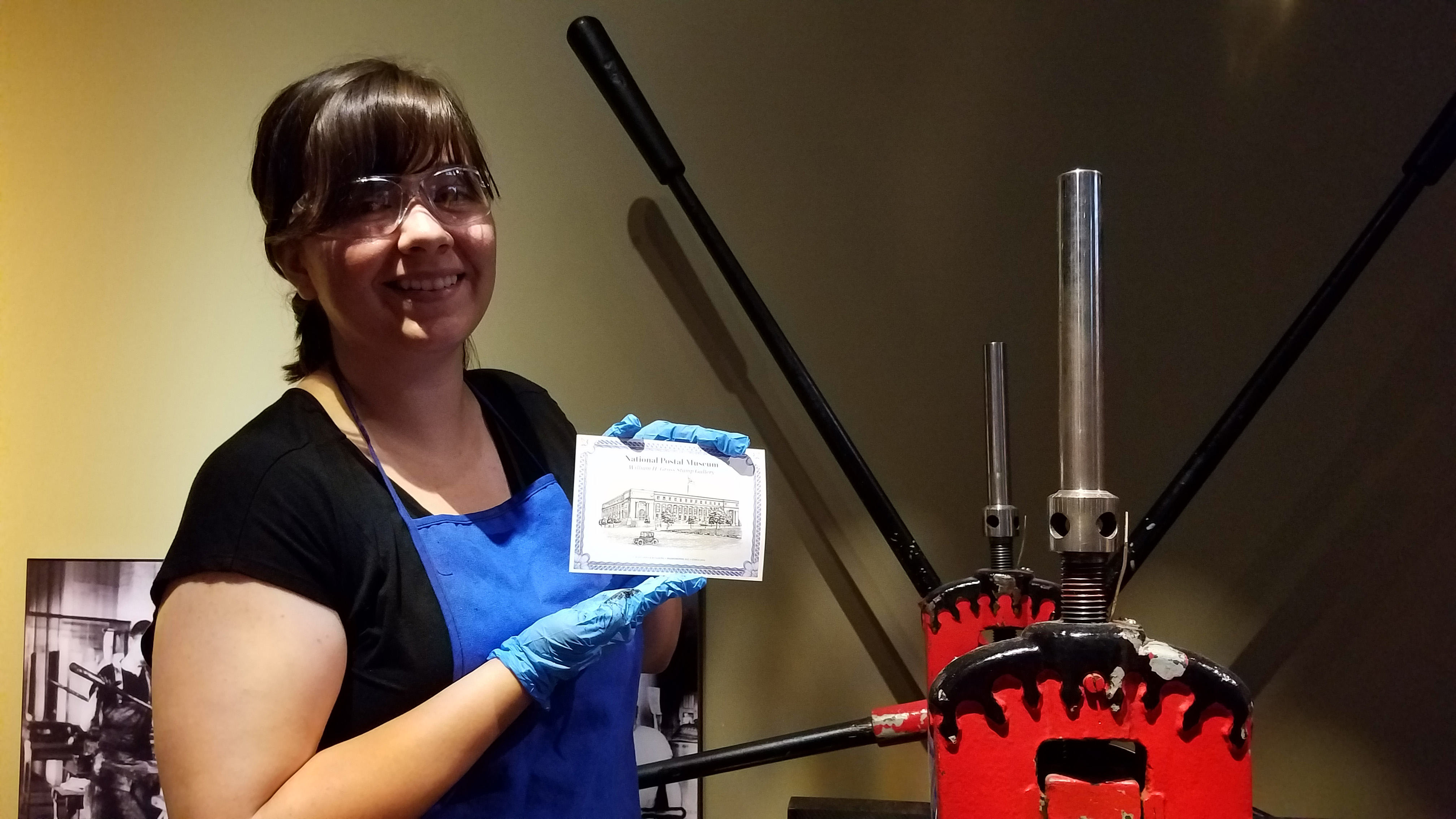 Woman wearing safety glasses, a blue apron and blue latex gloves, holding a postcard that she pressed using a machine with long levers.