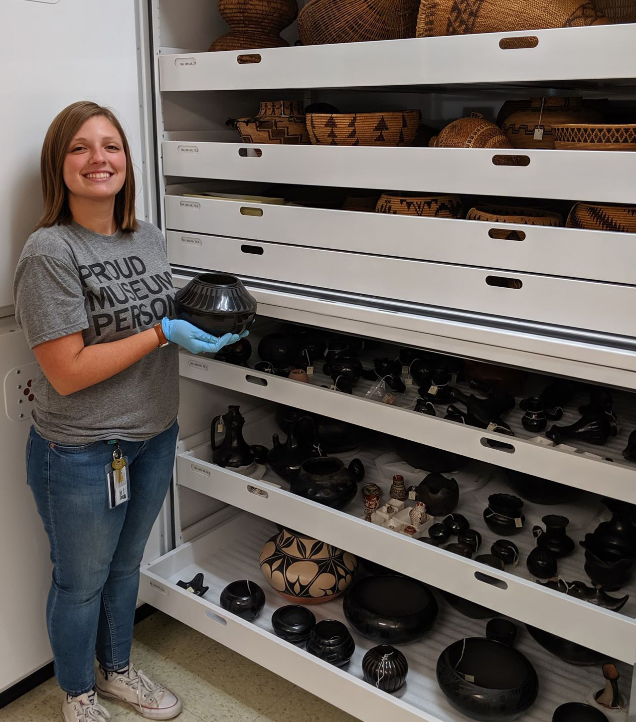 Woman standing in front of a large bank of collections drawers, some pulled out to display  pottery and baskets, while she holds a small black pot in her hands