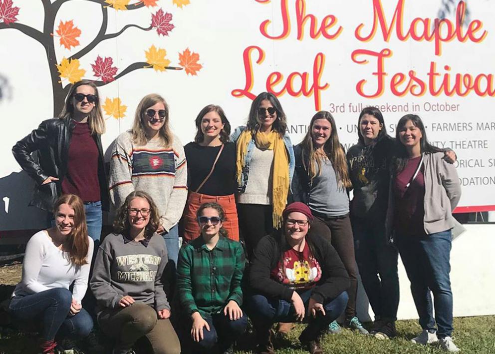Eleven women in front of the Maple Leaf Festival sign while smiling, with four of the women kneeling.